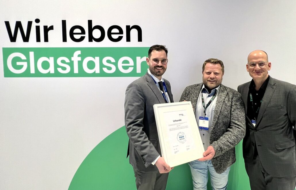 Drei Personen präsentieren stolz eine Urkunde vor dem Hintergrund des Slogans „Wir leben Glasfaser“ von Unsere Grüne Glasfaser.