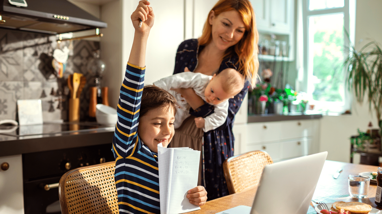 Glücklicher Junge zeigt seine Hausaufgaben an seine Mutter, während sie ein Baby im Arm hält, in einem modernen Küchenumfeld.