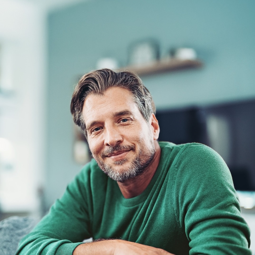 Mann in grünem Pullover lächelt zufrieden in modernem Wohnraum, symbolisiert die Verbindung mit Unsere Grüne Glasfaser.