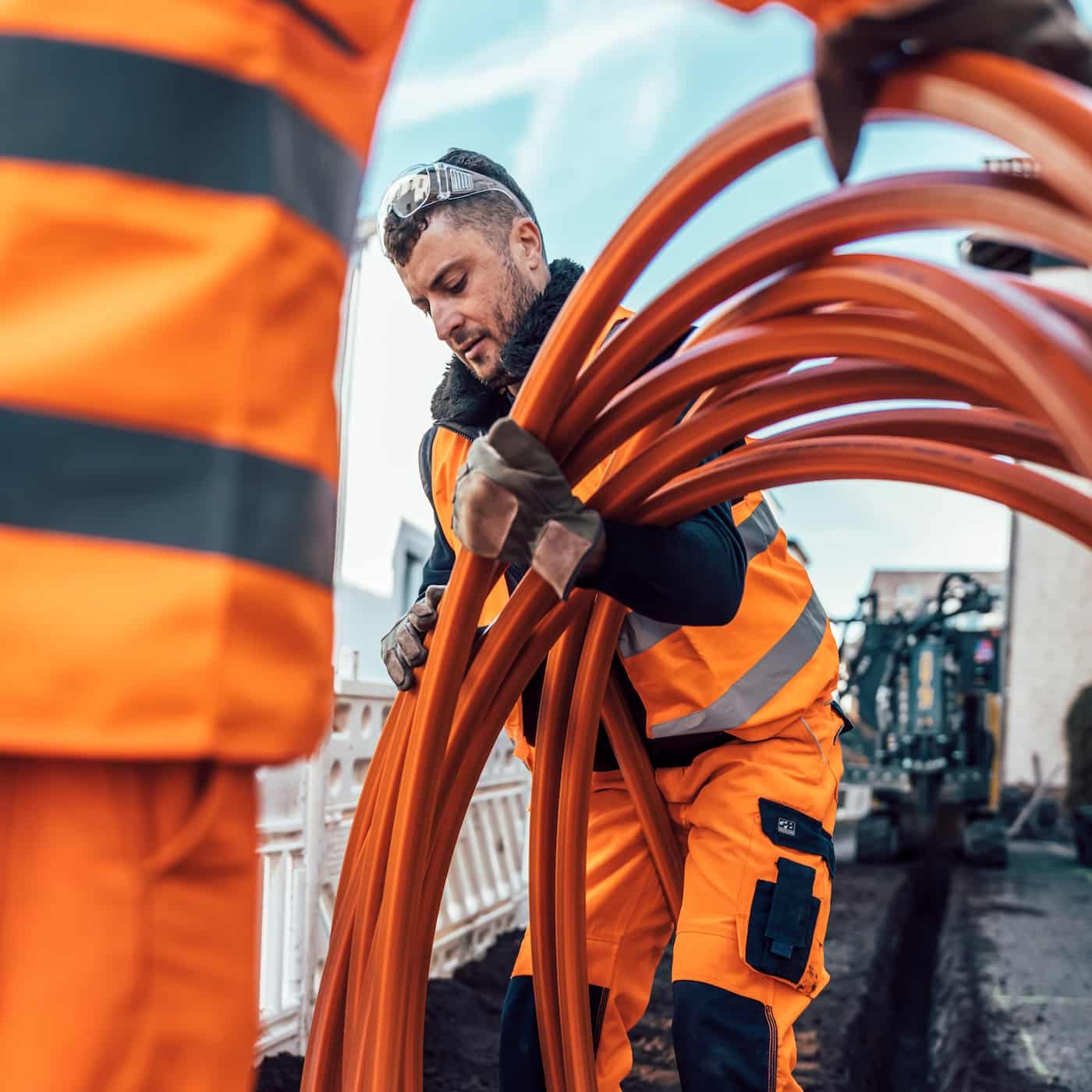 Fachkraft verlegt orangefarbene Glasfaserkabel auf einer Baustelle für Unsere Grüne Glasfaser. Effiziente Internetanbindung sichern.