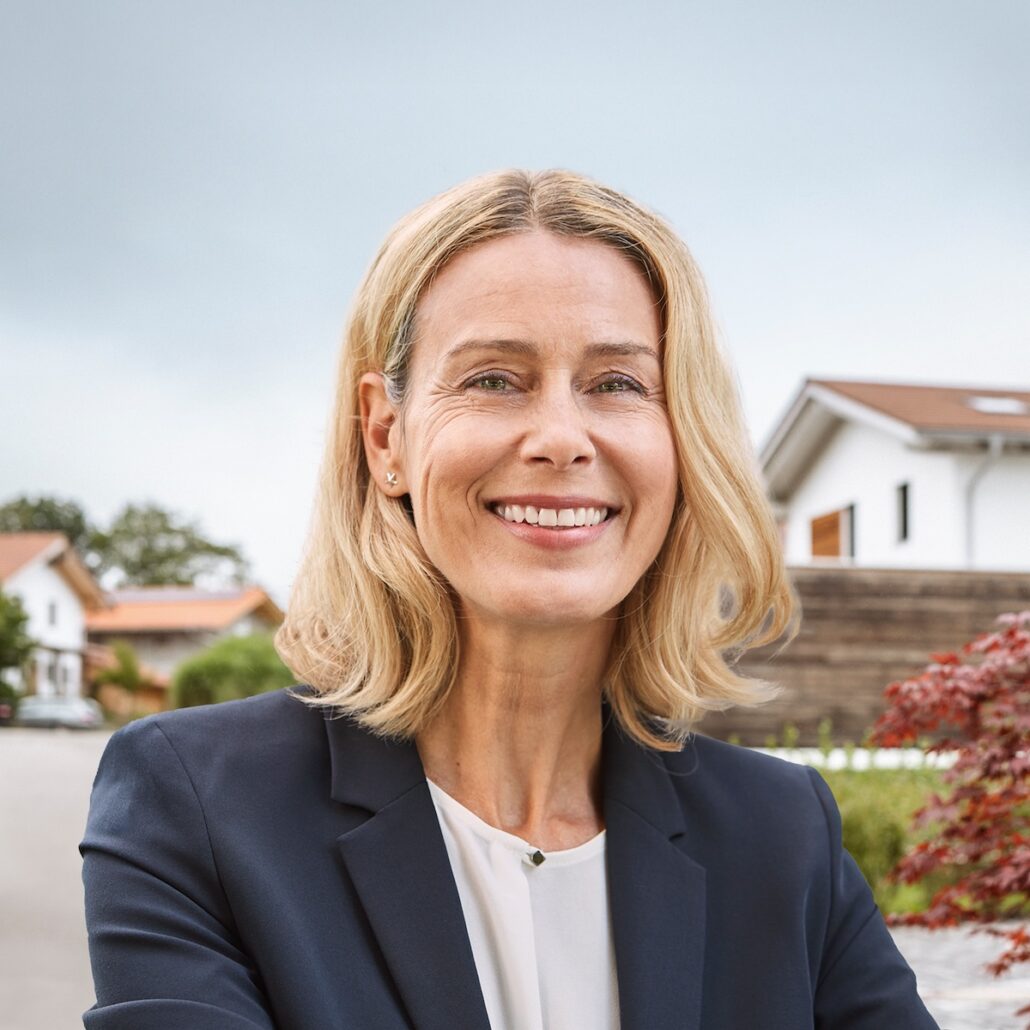 Frau mit blonden Haaren in Businesskleidung, lächelt vor einem Wohngebiet, symbolisiert die Zukunft von "Unsere Grüne Glasfaser".